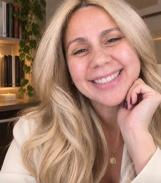 Luciana Lima sorrindo, com a mão apoiada no rosto, diante de uma estante com livros e uma planta.